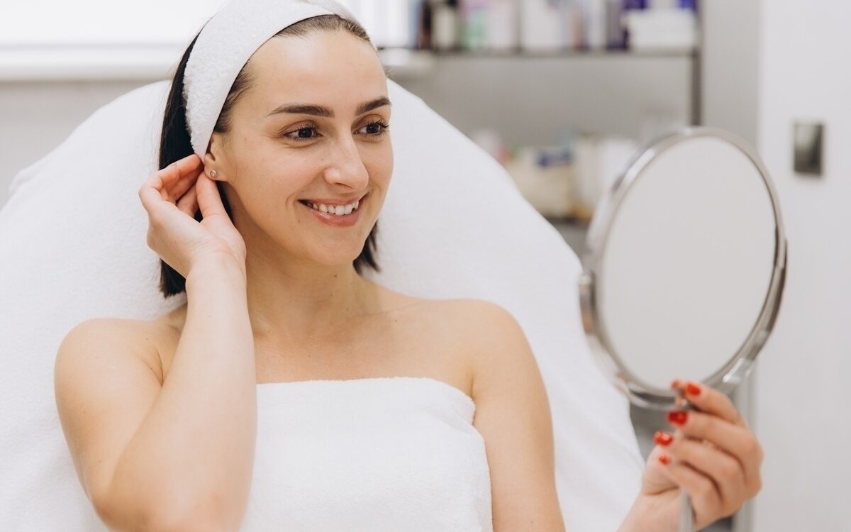 How Long Do Morpheus8 Results Actually Last? 9 Patient in a clinical setting holding a mirror and smiling at her smooth healthy skin reflecting visible Morpheus8 results after radiofrequency microneedling treatment
