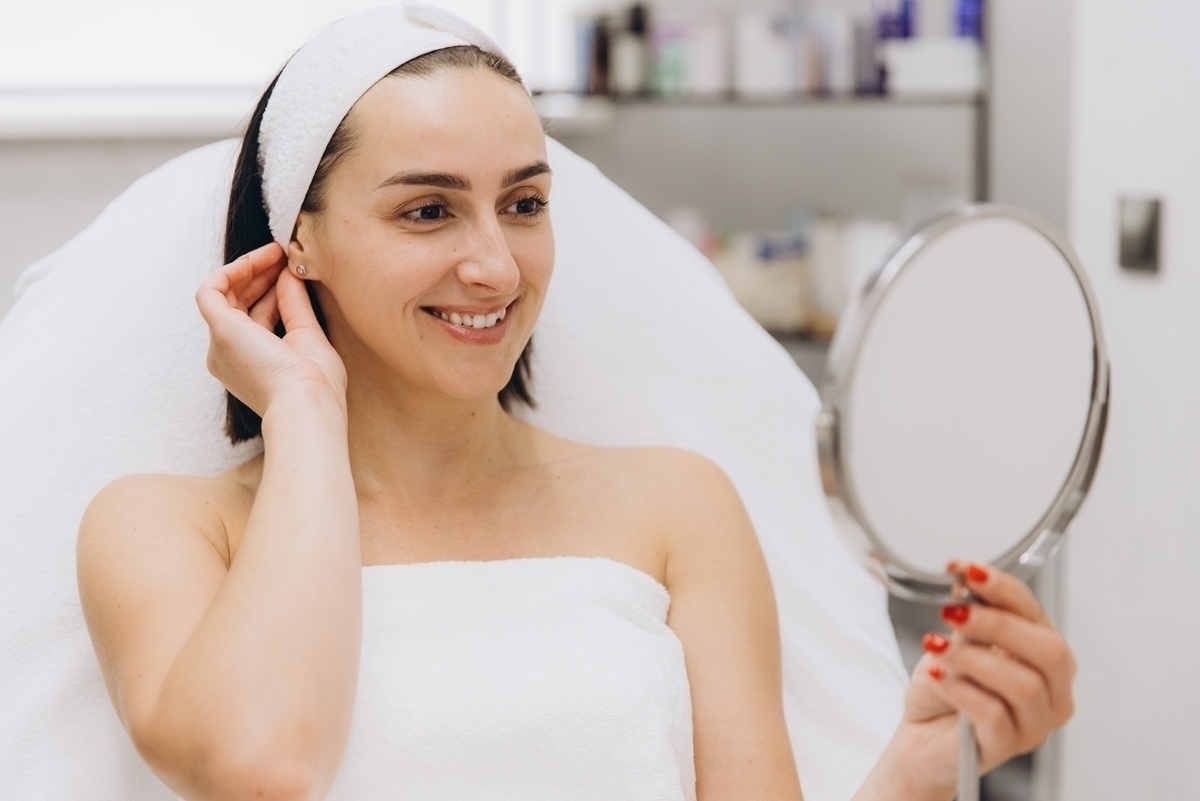 Patient in a clinical setting holding a mirror and smiling at her smooth healthy skin reflecting visible Morpheus8 results after radiofrequency microneedling treatment