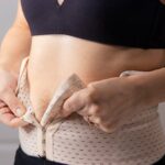 Woman fastening a postpartum compression garment around her abdomen with visible loose skin representing the body changes that Quantum RF post-pregnancy treatment addresses